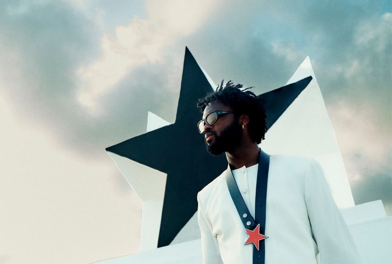 Musician Genesis Owusu wears sunglasses and a white jacket in front of a large black-and-white star sculpture against a cloudy sky.